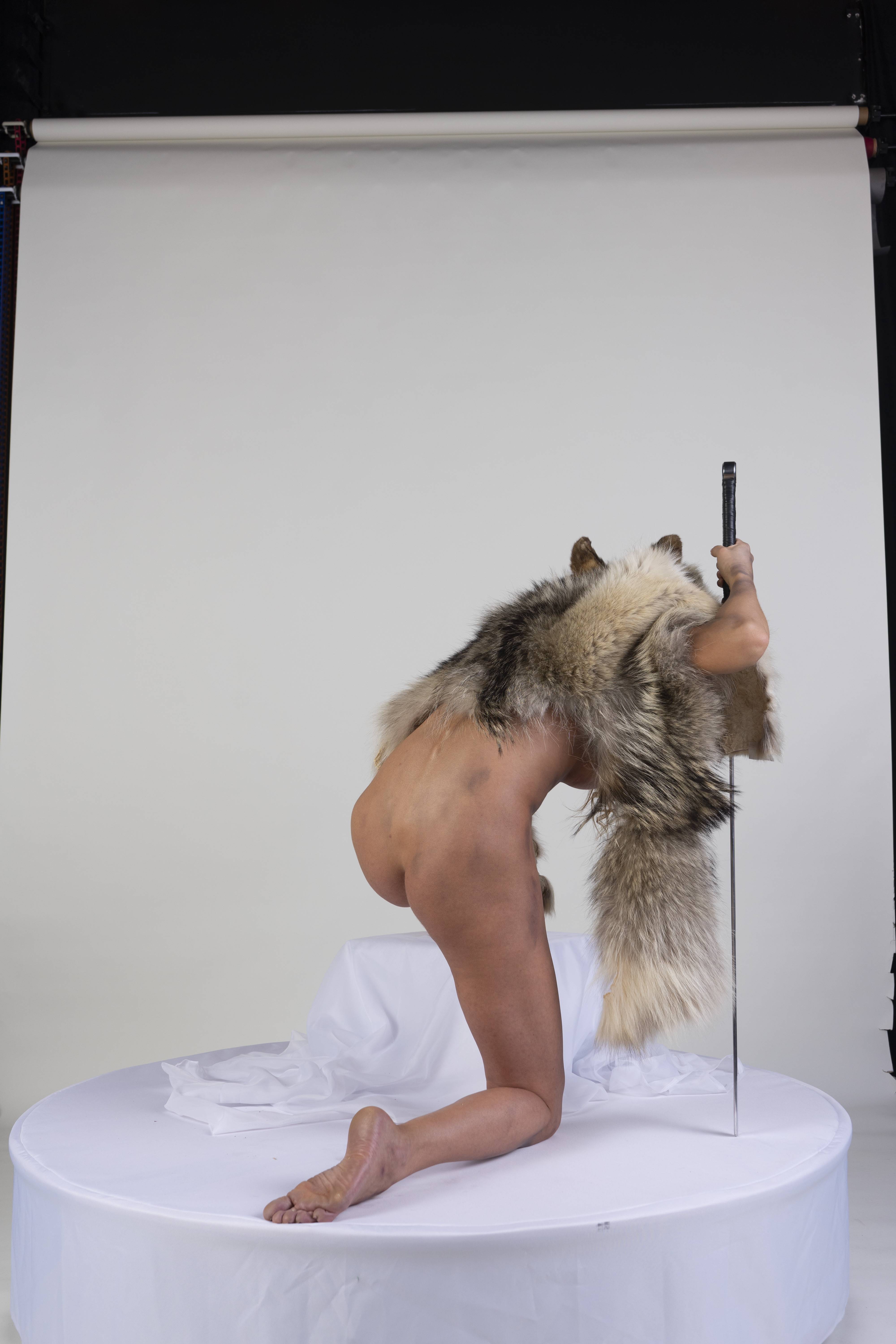 a nude woman kneels on a round platform covered with a white sheet in a studio setting. she is draped with a fur pelt and holds a pole. the background is a plain white studio backdrop.