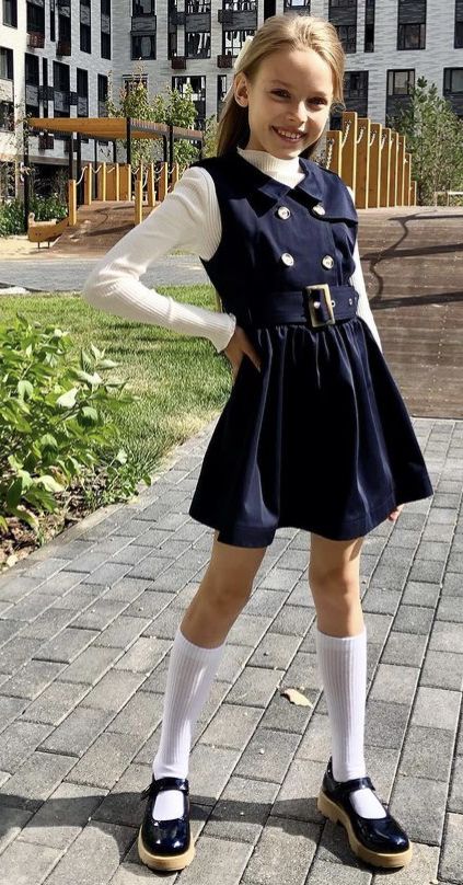 a fullbody shot of a young girl standing on a sidewalk in front of a modern apartment building. she's wearing a navy blue dress with a white collared shirt underneath, a matching vest, a brown belt, white kneehigh socks, and black mary jane shoes. her hair is styled down and she has a pleasant expression on her face. the overall style suggests a school uniform or a similar formal outfit. the background is slightly blurred but shows the building's exterior and a bit of landscaping.