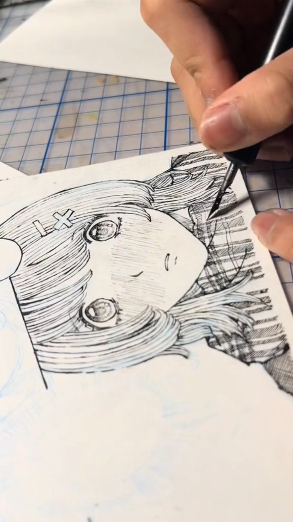 a hand drawing an anime style girl's face, hair, and shoulders on paper using a pen.  the artist is working at a table with a gridded surface.  the drawing is in progress, with only the girl's upper body drawn. the girl is looking to the right, has long hair, and is wearing a patterned shirt. the drawing is done in black ink on white paper. the artist's hand is clearly visible in the foreground of the picture.  the drawing surface is the focus of the image.