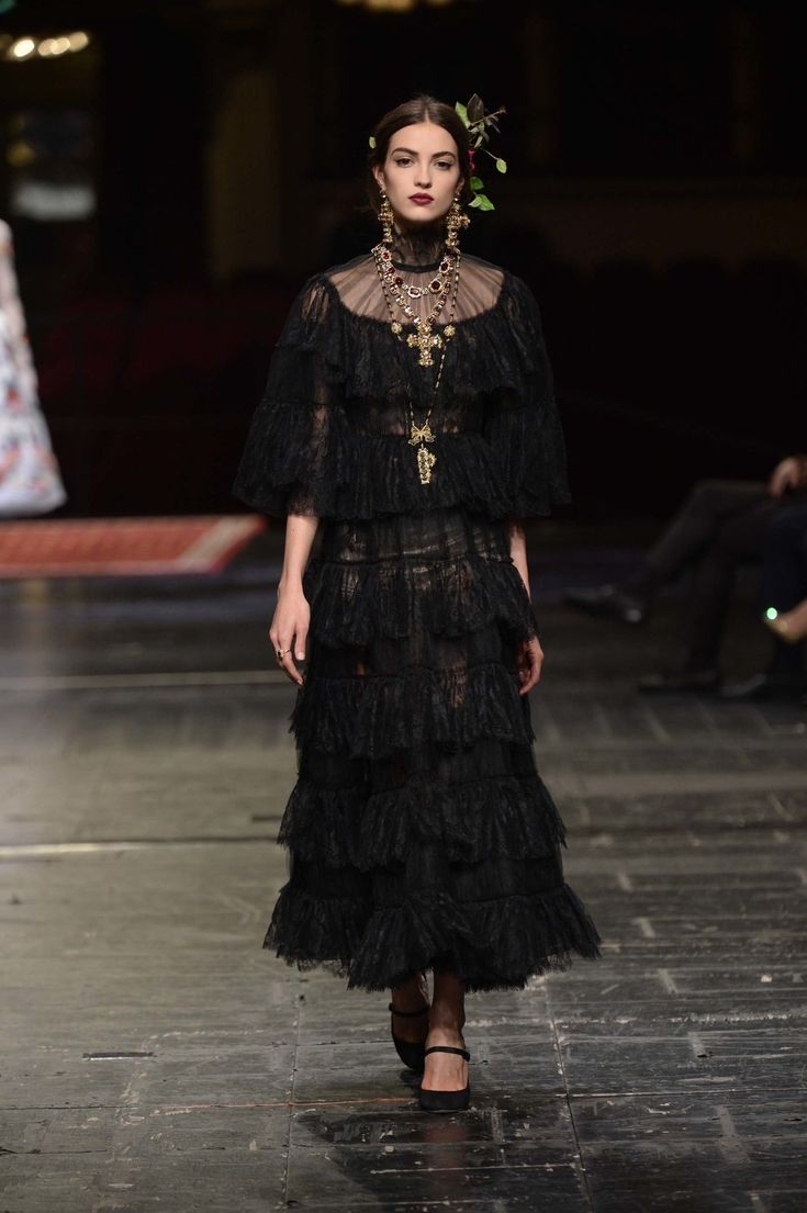 a model walks the runway wearing a stunning black lace dress. the dress features multiple tiers of ruffles and a high neck.  she is adorned with a gold necklace and earrings, and her hair is styled elegantly. the overall aesthetic is dark, sophisticated, and reminiscent of gothic fashion. the background is blurred, focusing attention on the model and her attire. the image is welllit and professionally taken, characteristic of a fashion show or editorial shoot.
