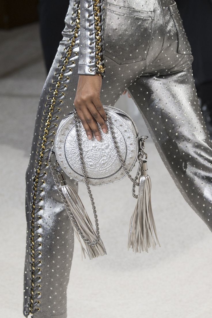 a woman wearing a silver outfit with chain detail. she is holding a silver round purse that is attached to a silver chain with tassels. the purse and outfit are adorned with silver embellishments. the woman is standing on a white surface.