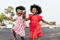 two young girls with big smiles are joyfully running on a dark sand beach. they are both wearing brightly colored dresses, one red and one floral. the girl on the right is holding a toy airplane in her hand. palm trees and other tropical foliage can be seen in the background.