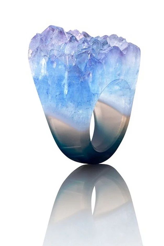 a close up of a unique agate ring. the ring is made of agate that is blue, purple, and white. the ring is shaped like a crystal and has a rough, natural look. the ring is shown on a white background and is reflected in the surface. the image shows a highquality product and looks elegant and stylish.