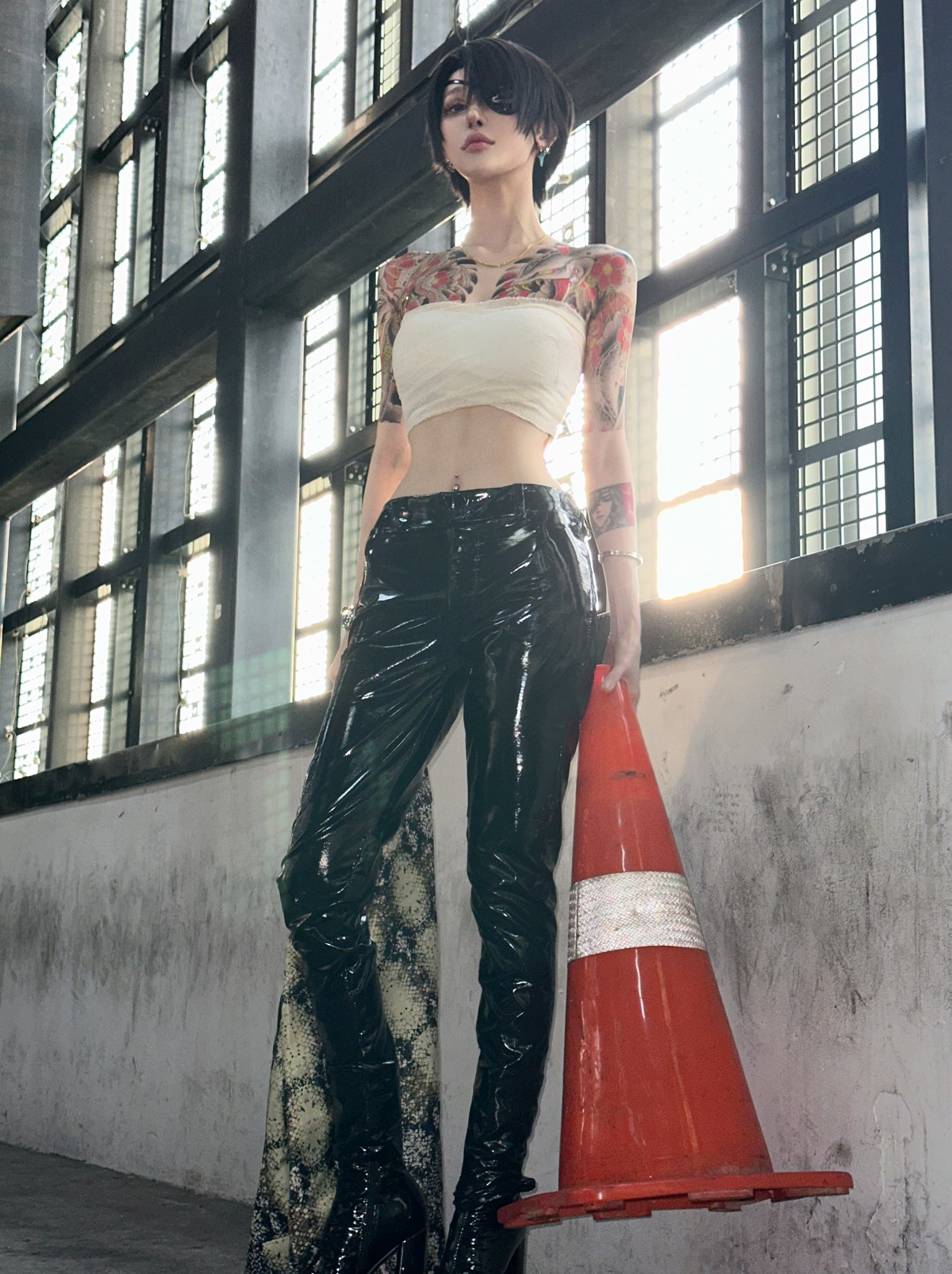 a woman in a cosplay outfit stands confidently, holding a traffic cone. she's wearing black leather pants, a white crop top, and high black boots. her arms are adorned with tattoos. the background is an urban industrial setting with large windows. the overall style is edgy and bold.