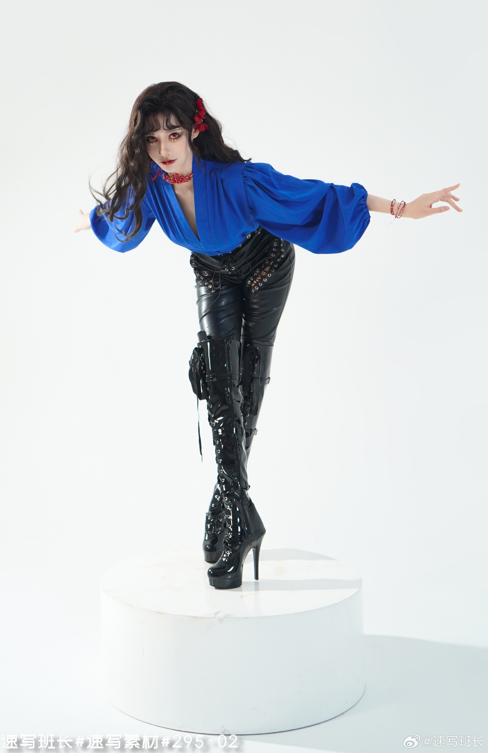 a woman in a blue blouse, black leather pants, and black platform boots stands on a white platform. she has curly black hair and red lipstick. the woman is posing with her arms outstretched. the background is white, and the lighting is studio lighting.