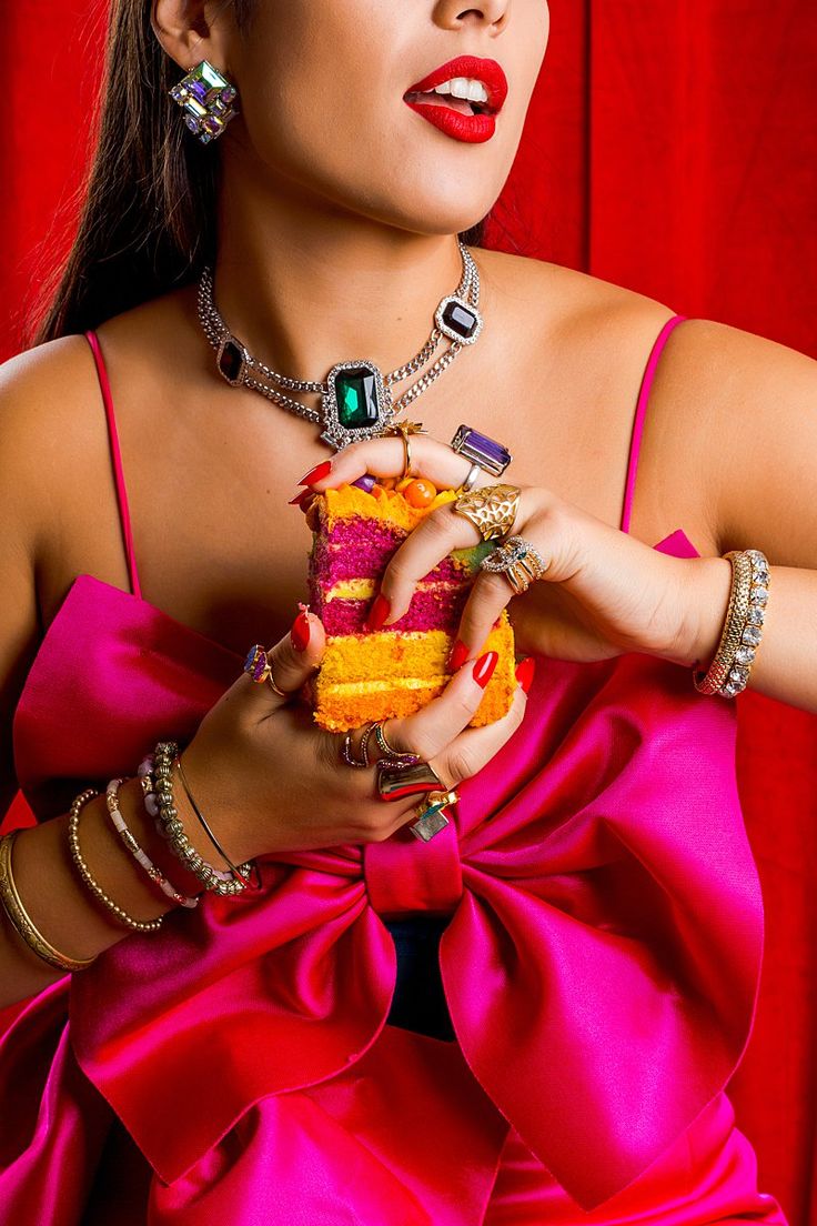 loseup view of a woman wearing a vibrant pink dress and an abundance of colorful jewelry. she is holding a multilayered cake, which is also richly colored.  the setting is against a dramatic red backdrop, creating a highly stylized and luxurious aesthetic. the focus is on the woman's bold style and the rich textures and colors in the image.