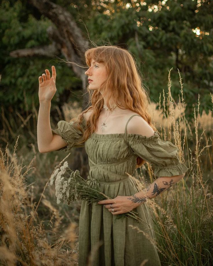 a young woman with long red hair is standing in a field of tall grass, looking up and smiling. she is wearing a green dress and has flowers in her hand. she is enjoying the summer day and looks happy and carefree. the image is warm and natural, with soft lighting and a relaxed, boho style.
