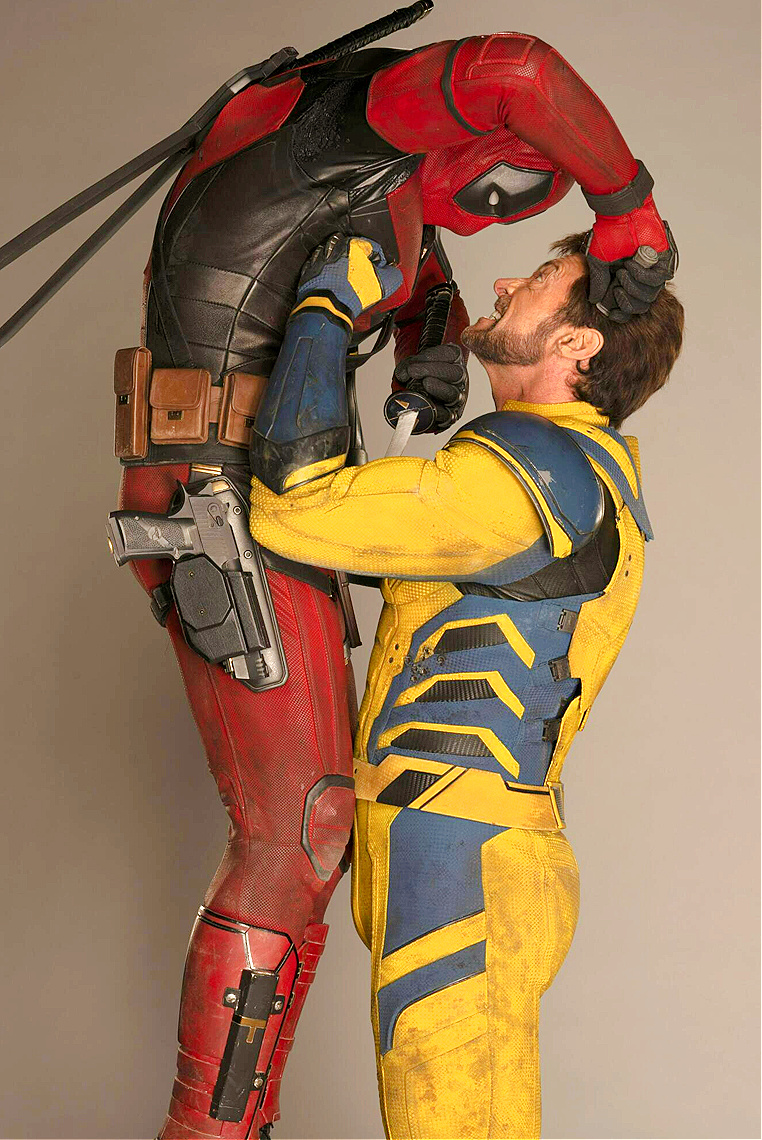 deadpool is seen holding wolverine by the neck with a knife to his throat. deadpool is wearing his red and black suit, and wolverine is wearing his yellow and blue costume. they are engaged in a mock fight or playful interaction.