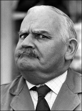 this is a black and white closeup portrait of an older man. he has a strong facial expression with raised eyebrows and a furrowed brow. his mouth is slightly open, creating a look of surprise or disapproval. he has a neatly trimmed mustache and is wearing a collared shirt and tie with a suit jacket. the background is blurry, focusing attention on the man's face.