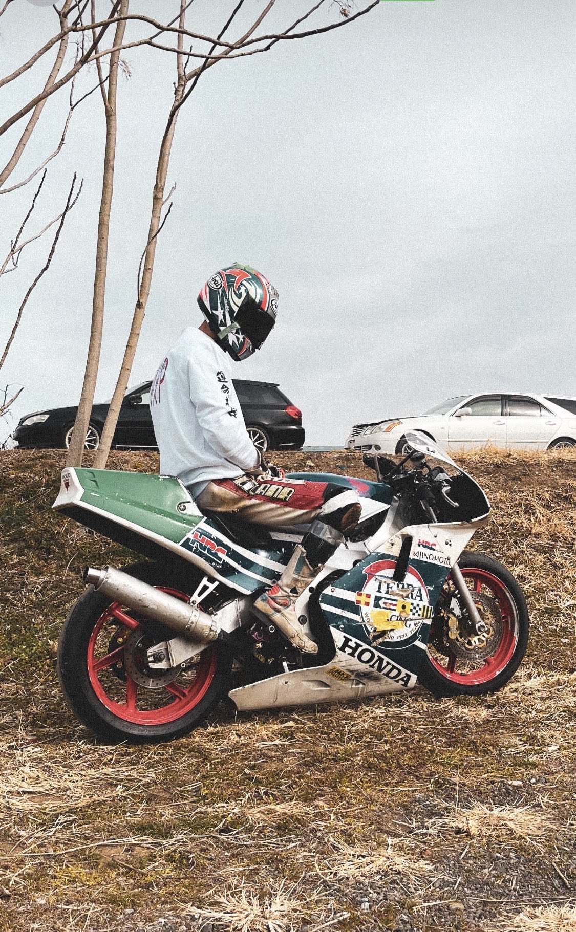 a man wearing a helmet is sitting on a honda motorcycle. it is a sunny day and the rider is dressed in racing gear. there is a car behind the motorcycle. the motorcycle is parked on the side of the road with dry grass in the foreground.