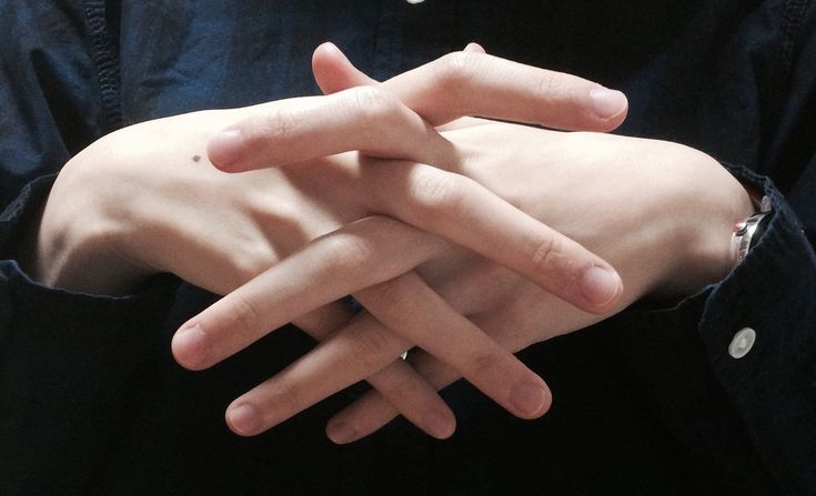 a closeup image of a man's hands, his fingers intertwined in a casual relaxed pose. his skin is pale and his nails are manicured. he is wearing a dark colored buttondown shirt. his left wrist is adorned with a watch.  the image is  minimalist and simple, capturing the beauty of a natural gesture.