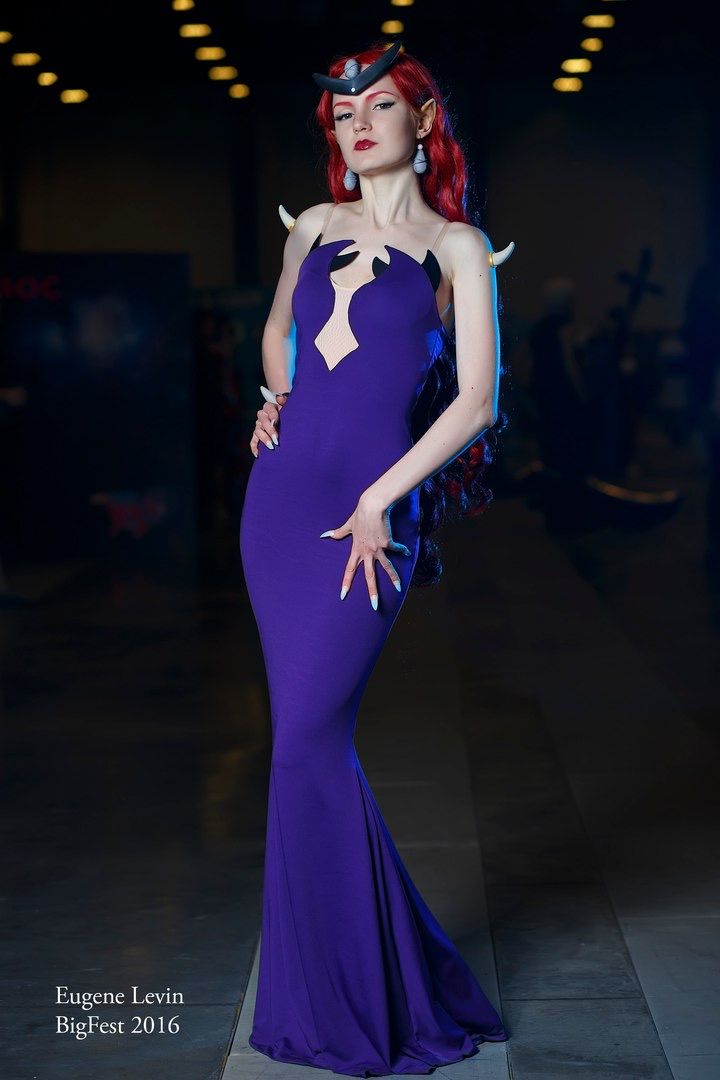 a woman dressed in a purple gown and costume, posing for the camera. the woman has elflike ears, red hair and wears a horn headdress. her dress has a front cutout and she has jewelry on.