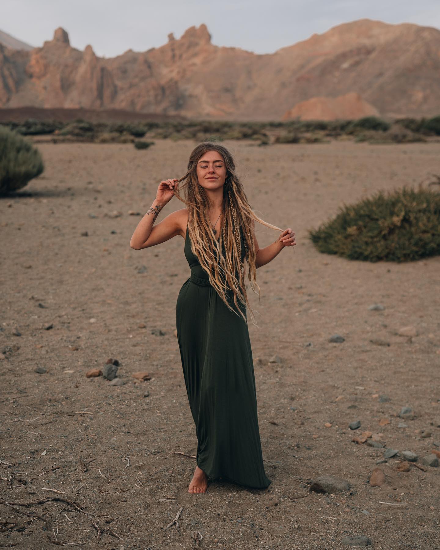 a woman with long blonde dreadlocks wears a dark green maxi dress. she stands barefoot in a desert landscape with mountains in the background.  the woman appears peaceful and relaxed, with her eyes closed, suggesting a sense of serenity and connection with nature. the setting appears to be a volcanic or arid region, possibly in the canary islands. the overall mood is bohemian and freespirited.