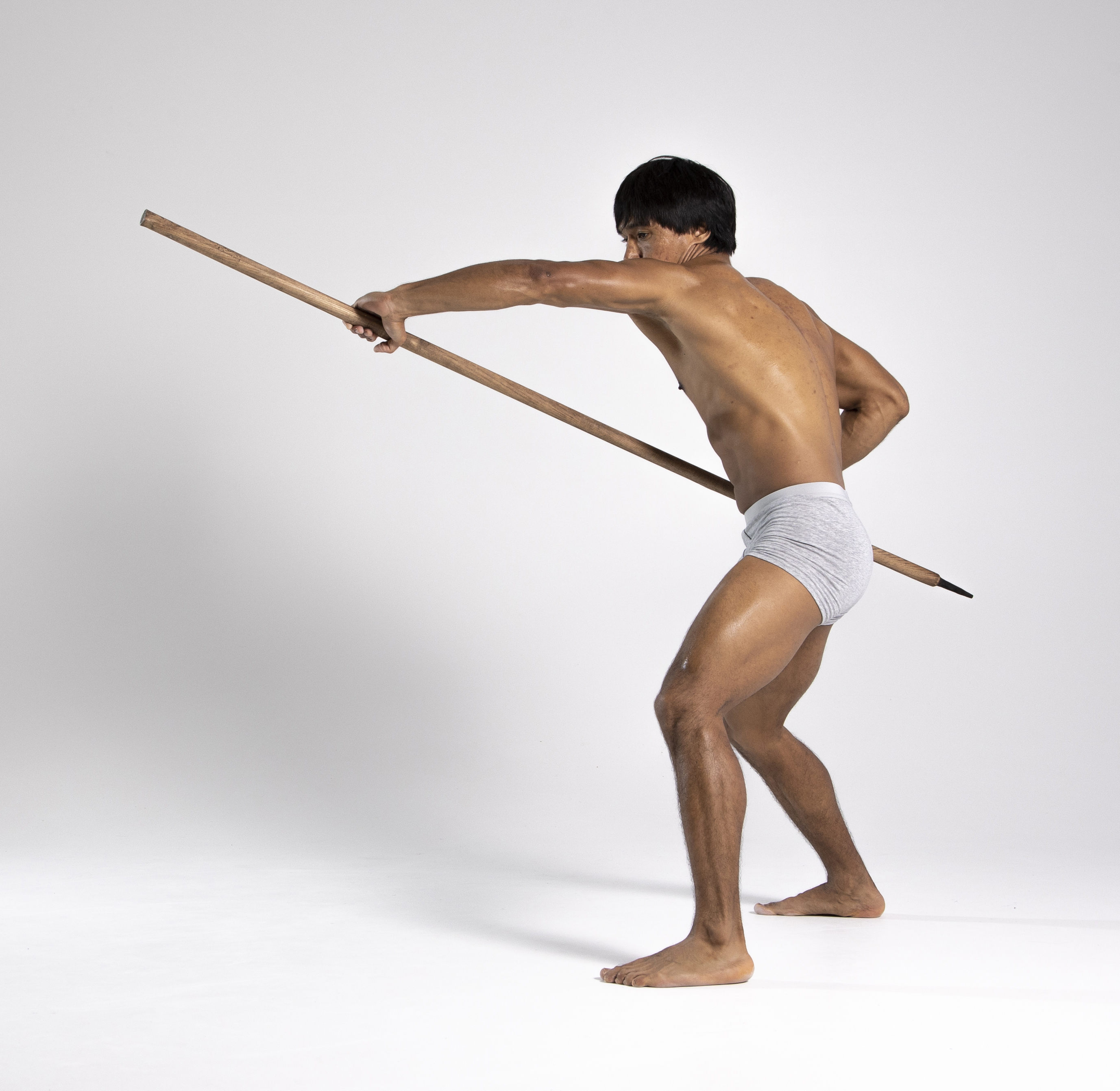 a shirtless man in gray underwear is holding a spear with both hands and is in a lunging pose. he is barefoot and standing in a studio with a white background.
