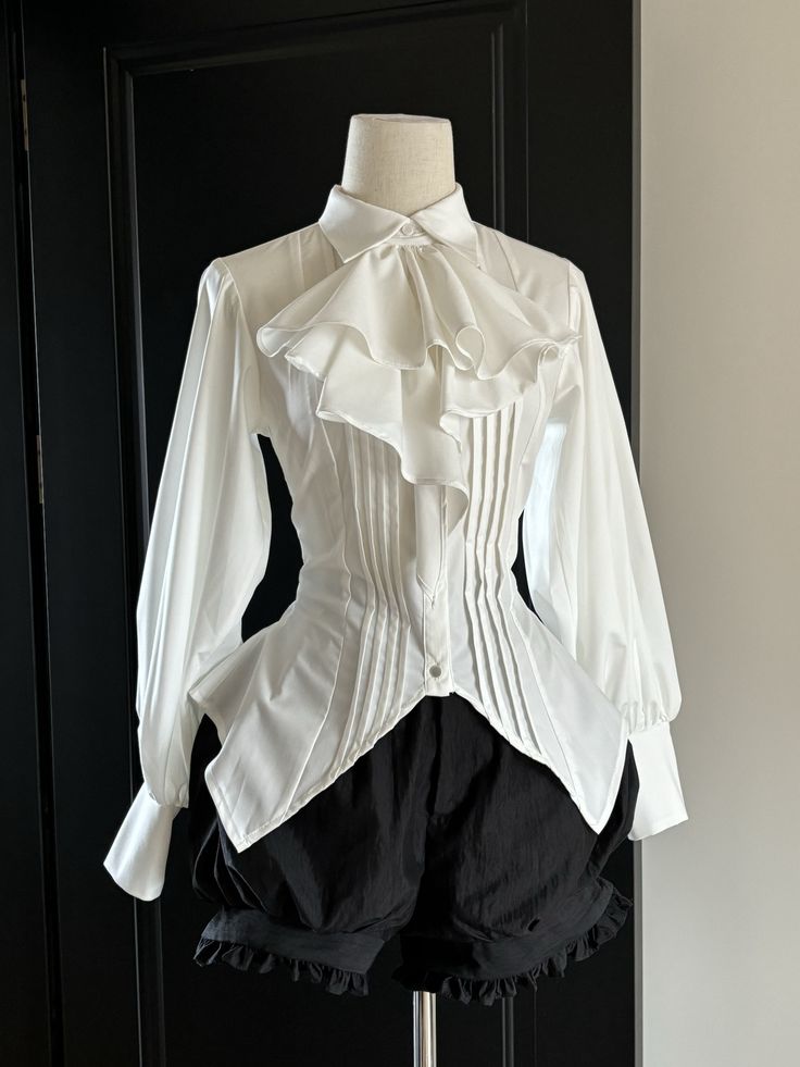 a white victorianinspired blouse with a ruffled collar and pleated bodice is displayed on a mannequin. the blouse has bishop sleeves and a highlow hem. it is paired with black bloomers. the background is a black door.