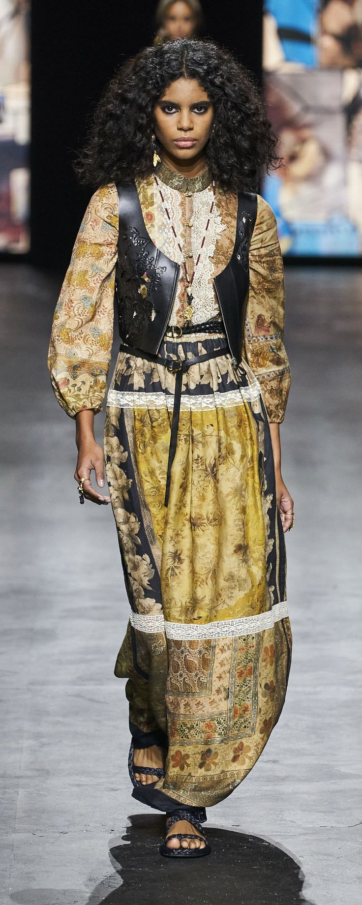 a fashion model walks the runway wearing a long, flowy dress with a vest, sandals, and jewelry. her hair is long and curly, and she has on makeup. the dress is patterned with flowers and has a long skirt.  she is on a runway.
