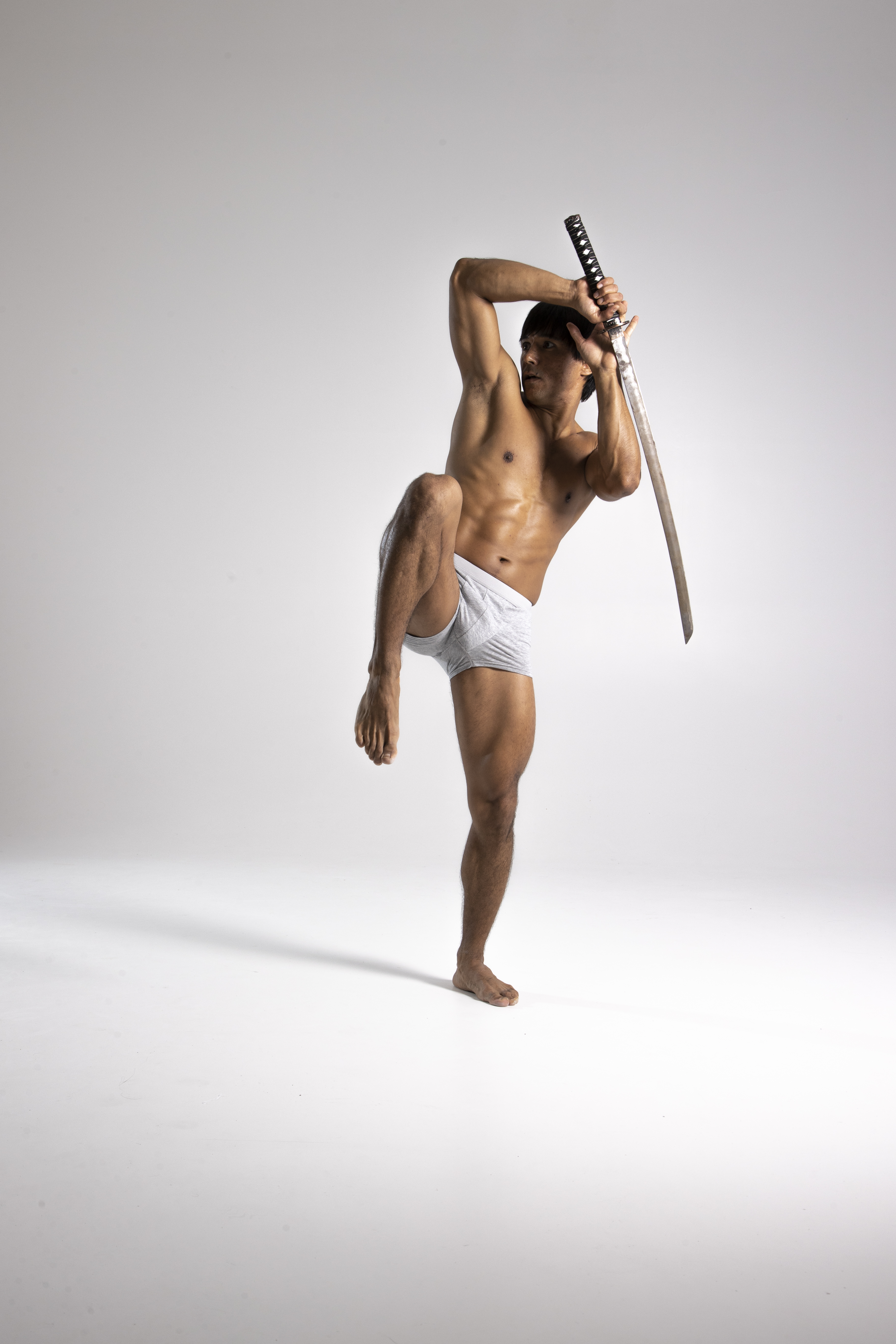 a shirtless man in white underwear poses in a warrior stance, holding a katana sword. he is athletic and muscular, and his body is facing forward while his head is turned slightly to the left. his left leg is raised with his knee bent in front of him. the background is plain white.