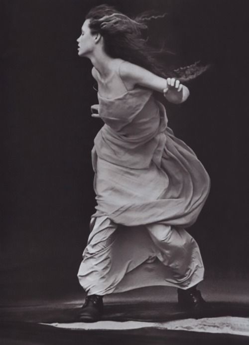 a woman in a long flowing dress is midstep with her long hair flowing in the wind.  the photo is in black and white.  the woman is turned to the side so we can see the side of her face and the back of her body.