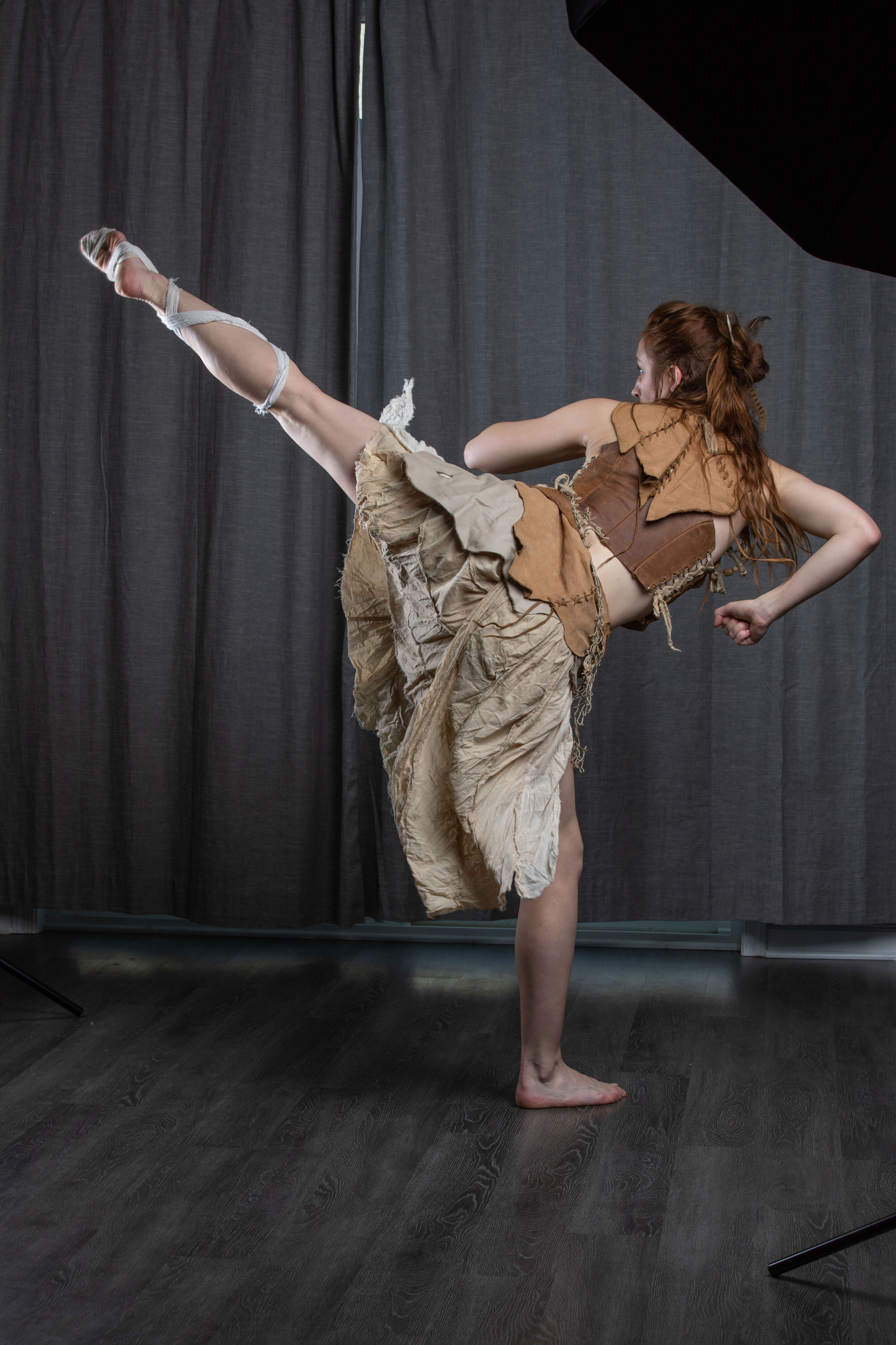 a woman in a warriorinspired costume performs a high kick against a dark studio backdrop. she is barefoot and wears a tattered, flowing skirt and a leather bodice. the lighting is dramatic, highlighting her form and the movement of her clothing.