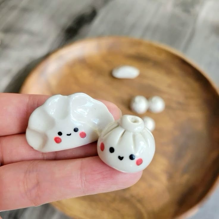 closeup shot of a hand holding a pair of kawaii ceramic food figurines, one a dumpling and the other a bun, each adorned with a cute face. a wooden plate with more food figurines is in the background.