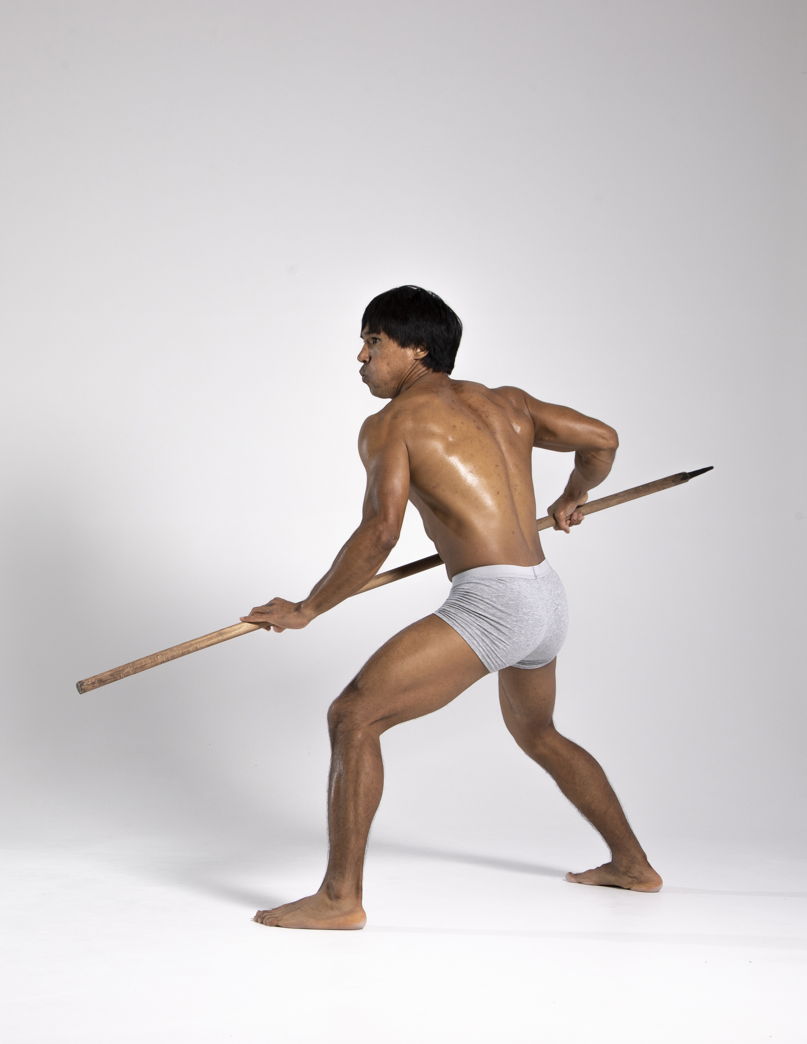 a man is standing in a studio, holding a spear. he is wearing underwear and is barefoot. he is looking over his shoulder. the photo is taken from a back view.