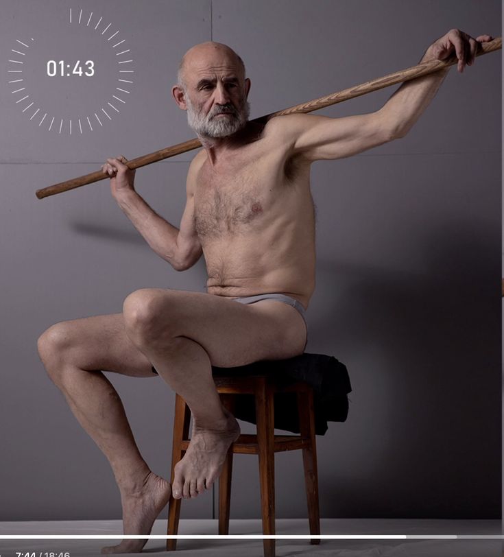 the image shows an elderly, nude male model sitting on a wooden stool. he's holding a long wooden pole across his shoulders.  his body is angled, and his expression is serious. the background is a plain grey, and a timer showing "01:43" is superimposed in the top left corner. the overall impression is that this is a photograph taken for artistic purposes, likely for life drawing or anatomical study.