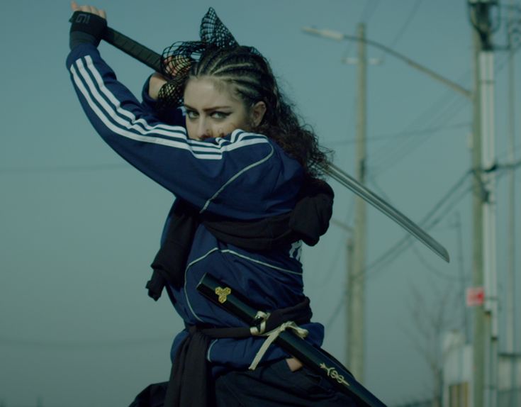 a woman in a blue adidas tracksuit and a black hood stands on a city street holding a katana sword, she is looking towards the right side of the frame. her eyes are intense, and she looks like a fierce warrior. the background is a blur of grey and green, giving the image a sense of motion and energy. the woman's pose and the blade of her sword are both sharp, making her look like a dangerous force. the image is powerful and dramatic, capturing the spirit of a true samurai.