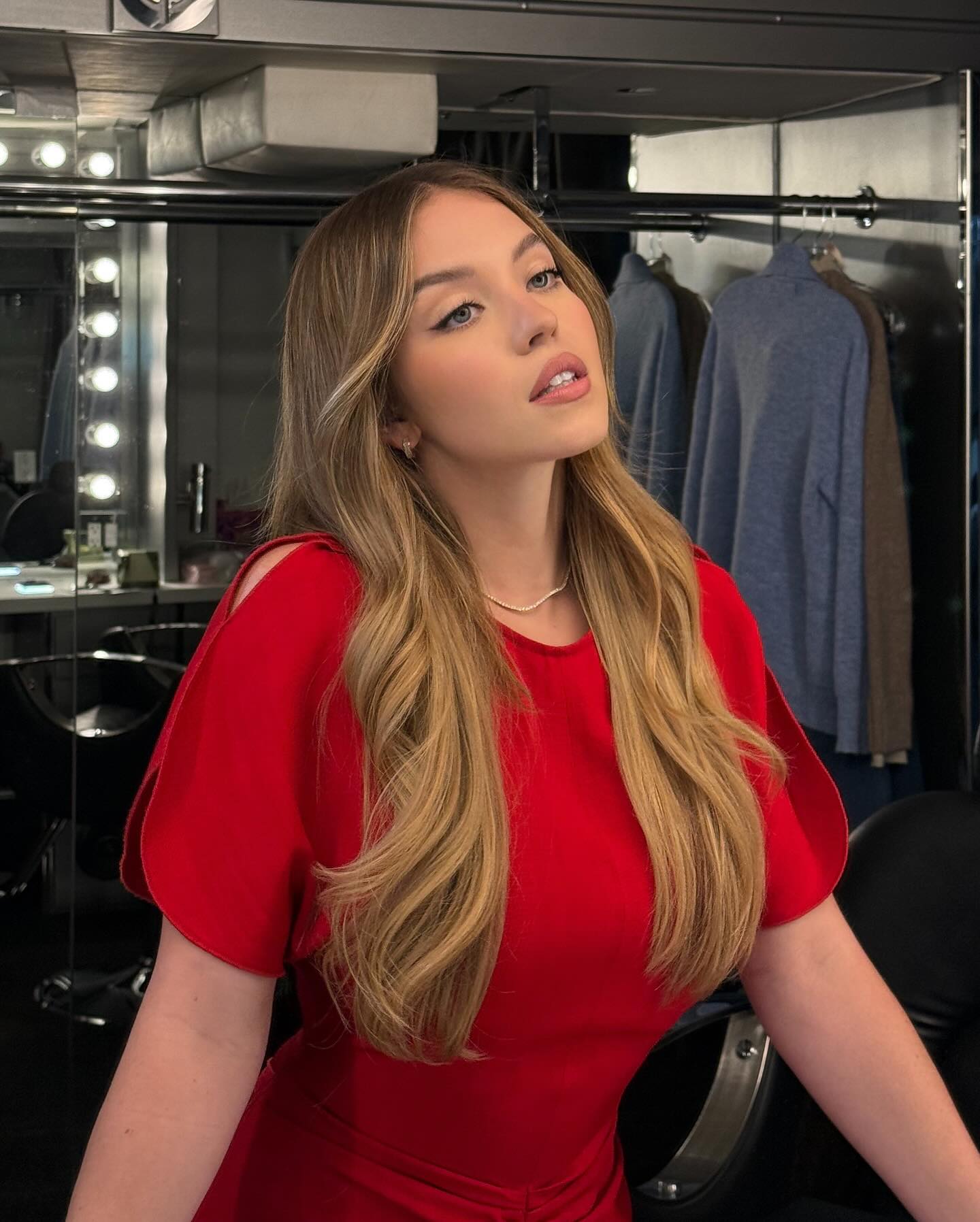 the image is a portrait of a young woman with long, wavy blonde hair. she's wearing a shortsleeved red dress with subtle shoulder cutouts.  the dress appears fitted. she has on makeup, including what seems to be winged eyeliner. the setting looks like a dressing room or backstage area, with a mirror and clothing racks visible in the background. the woman's gaze is directed slightly upward and away from the camera, giving a pensive or thoughtful expression. the lighting is focused on her, highlighting her features and the red dress.