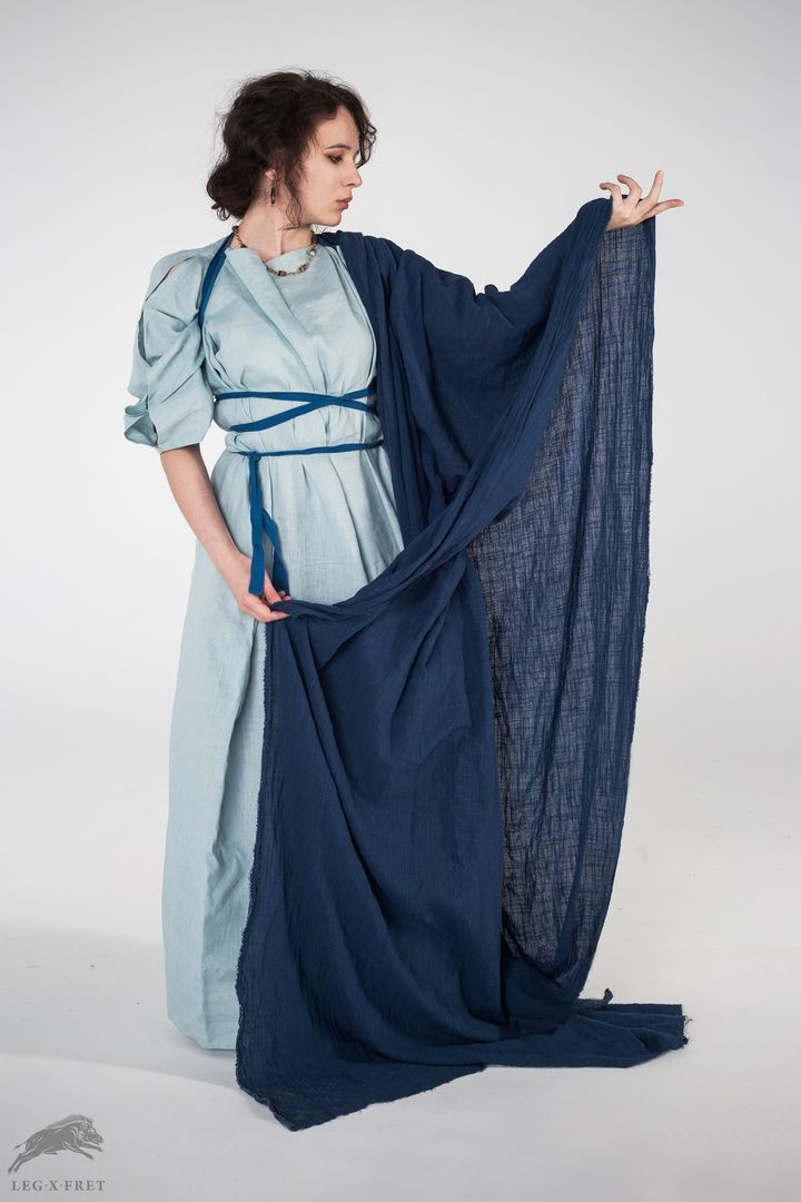 a woman in a light blue tunic and dark blue shawl, photographed in a studio setting. she is holding the shawl with her left hand and lifting it with her right, showing its texture. the tunic has a darker blue rope belt. the overall impression is that she is wearing an ancient or fantasy costume.