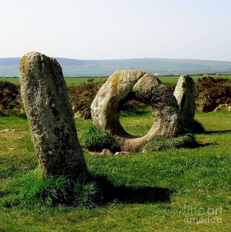 the image shows the menantol stone circle in cornwall, uk. it features a circular stone with a hole in the middle, flanked by two upright standing stones in a grassy field. the background includes a landscape of rolling hills and a clear sky.