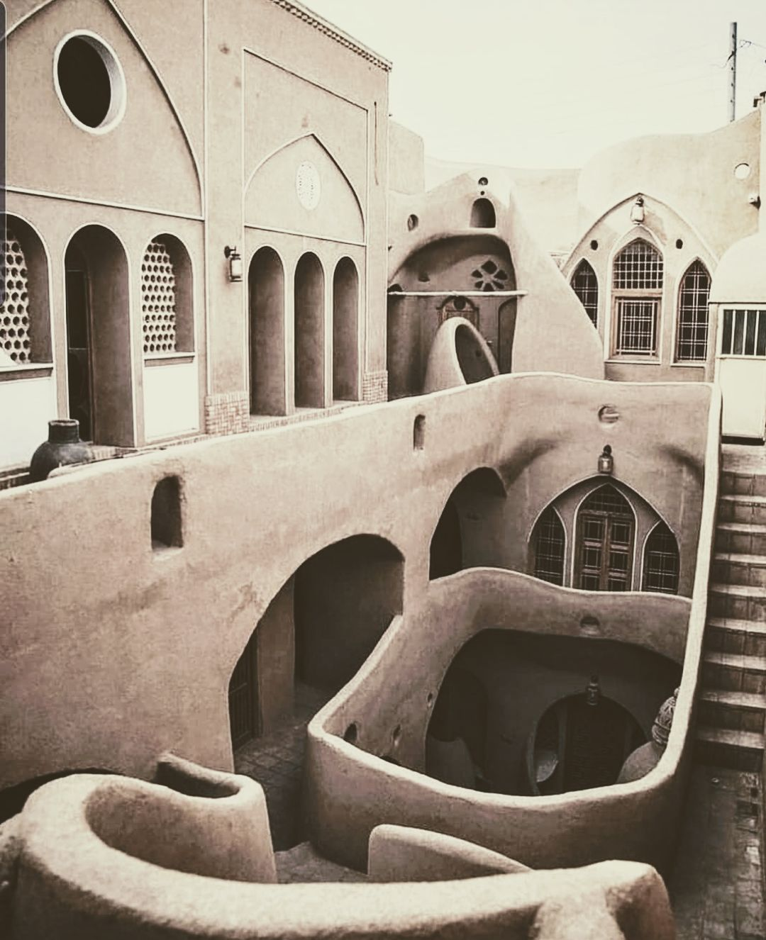 this sepia toned image captures an architectural scene. the composition features interconnected buildings and structures with recurring arches, creating a sense of depth and complexity. stairs lead to different levels within the courtyard, and windows punctuate the facades of the structures. the architecture appears to be in a middle eastern or islamic style, with clay or mud brick construction. the monochrome palette and the sepia tone create a vintage, timeless atmosphere, highlighting the architectural details and textures.