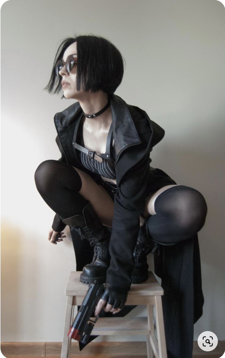 a young woman in a black leather jacket and black tights poses with a gun while sitting on a stool. she's wearing dark sunglasses, a black choker, and black boots. the background is a white wall. she has short black hair and her face is serious.  the photo has a mysterious and edgy vibe.