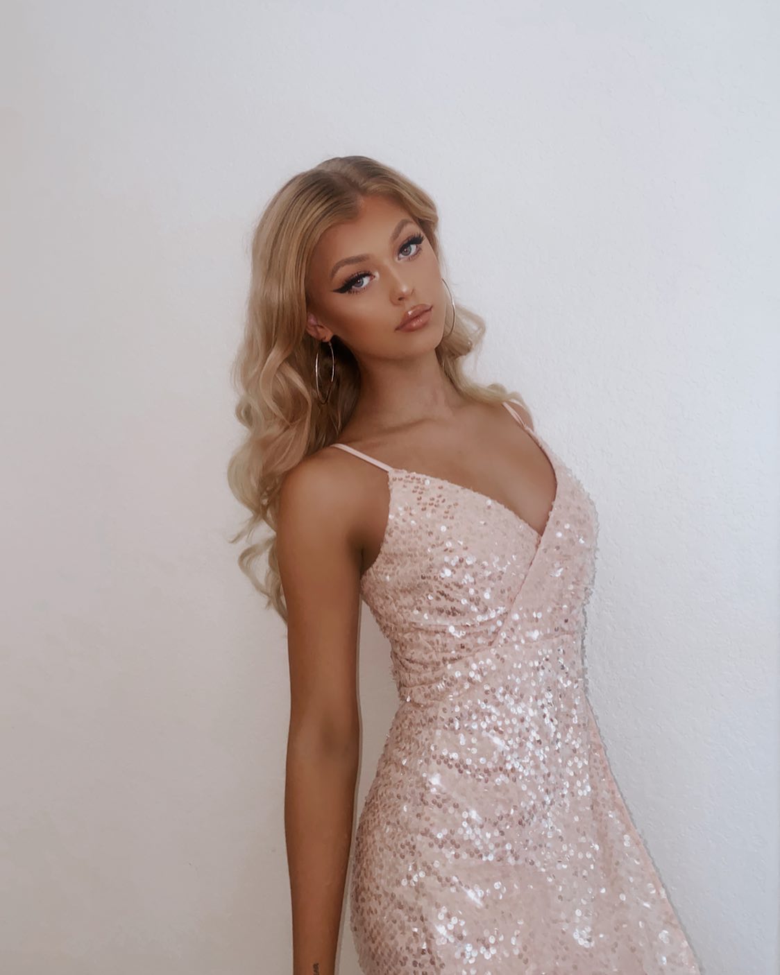 a fullbody shot of loren gray, a young woman with long, wavy blonde hair, wearing a light pink sequin spaghetti strap dress and hoop earrings, standing against a white background. she is posing and looking at the camera with a slightly tilted head. she has tan skin, and is wearing eyeliner and lipstick.