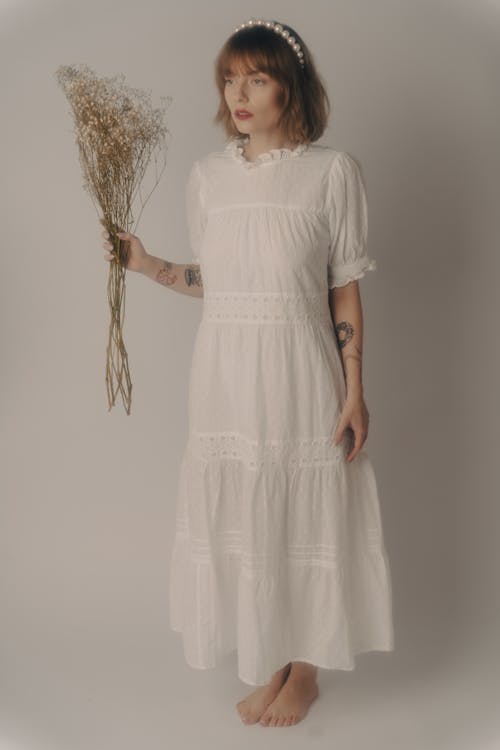 a young woman with short brown hair wearing a pearl headband stands barefoot in a white, tiered cotton dress. she holds a bouquet of baby's breath in her left hand. the background is a plain light grey studio backdrop. the overall style is romantic and elegant.