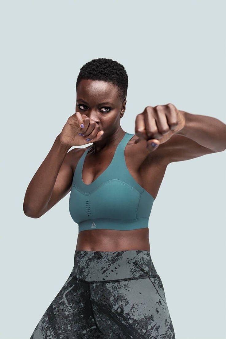 a black woman wearing a blue sports bra and grey leggings throws a punch.  she is looking towards the camera with a determined expression.  the background is light blue.