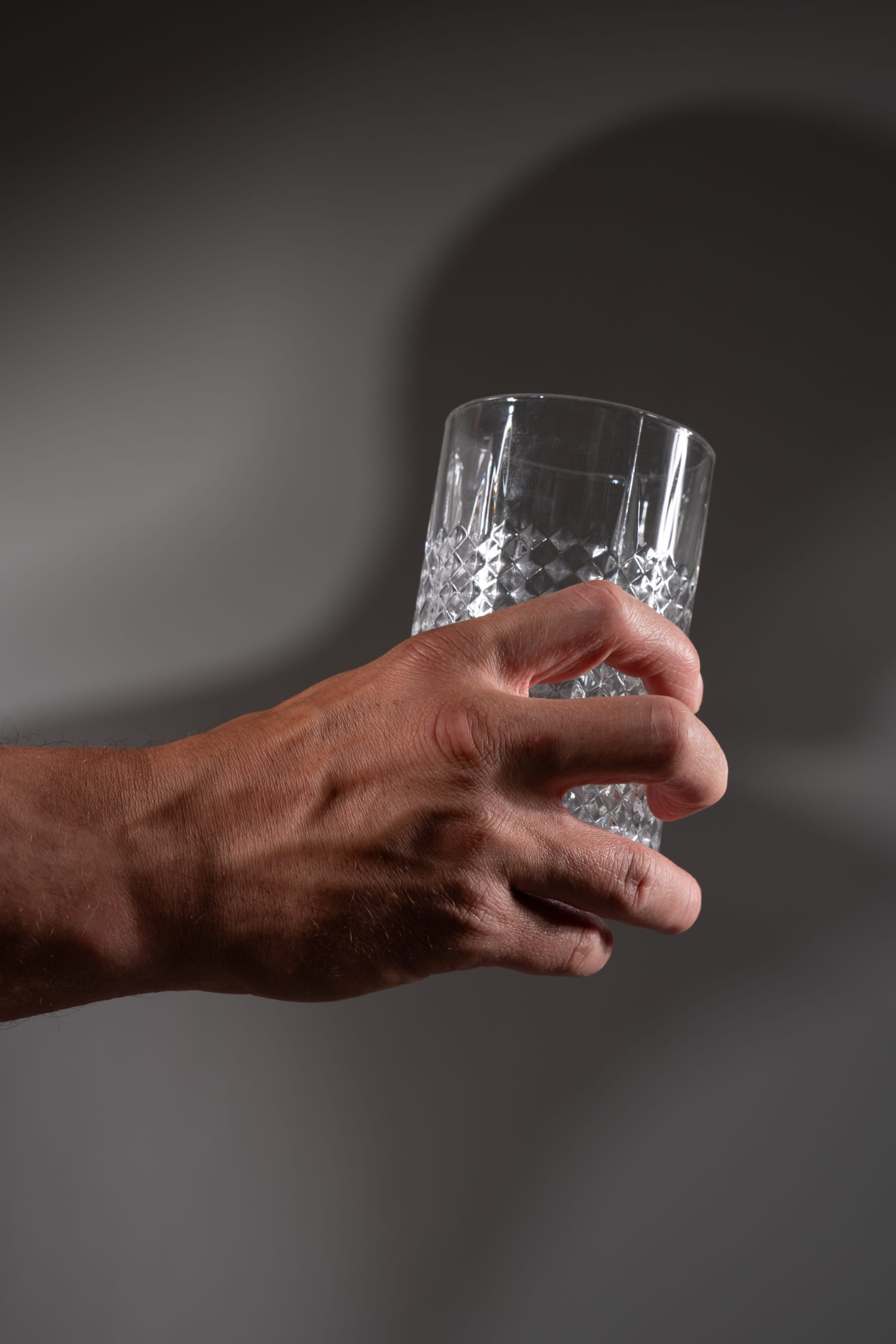 a man's hand holds a clear, empty, textured glass against a gray background. there's a distinct shadow of the hand and glass cast upon the background. the image is welllit, likely by studio lighting.