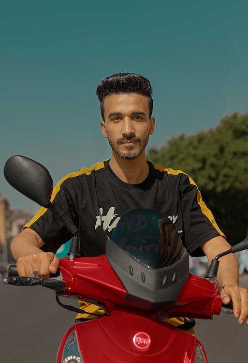 a young man with short dark hair and a beard sits on a red scooter, looking directly at the camera. he is wearing a black tshirt with yellow accents on the sleeves. the background is a sunny urban setting with trees visible. the overall tone is bright and vibrant.