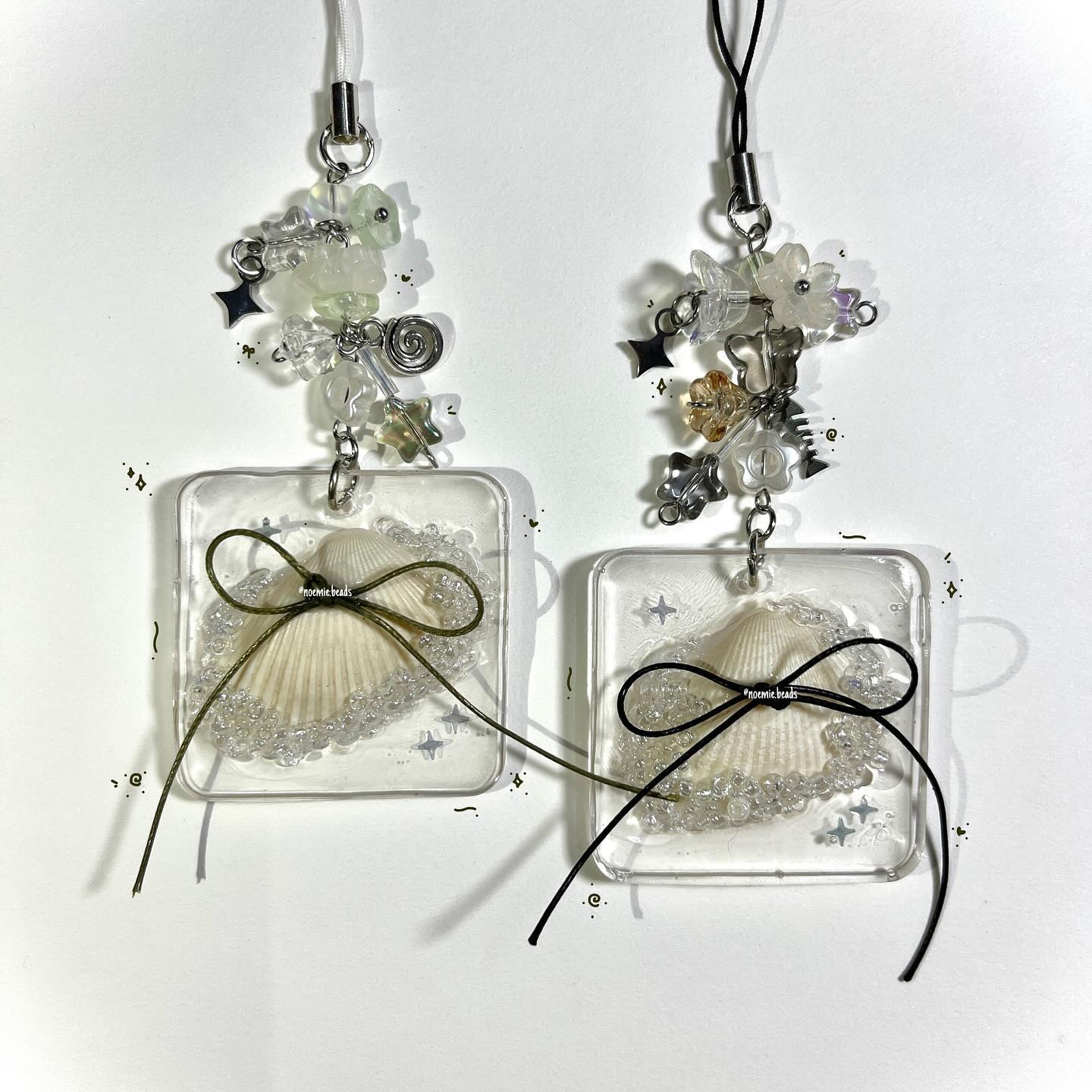 two seashell charms are displayed on a white surface. the charms consist of transparent square resin pieces housing a seashell surrounded by tiny white beads and tied with either a green or black bow. each charm hangs from a beaded strap adorned with flowers, stars, and various beads in white, green, silver, and amber. the charms are attached to the beaded straps with metal rings.