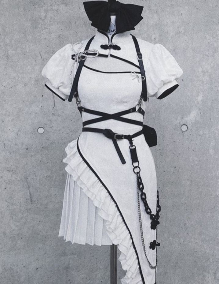 a white dress with black straps and a chain belt is displayed on a mannequin in a studio setting against a grey background. the dress has a ruffled hem and a high neck. the mannequin is wearing a black bow on its head. the image is high quality and welllit, showing off the details of the clothing.