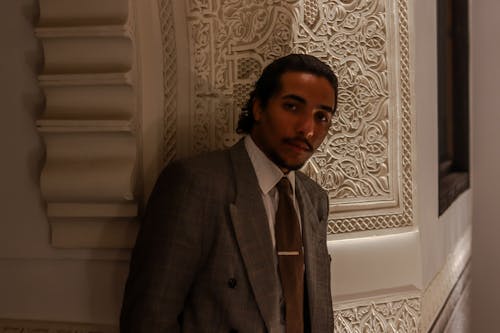 the image is a closeup portrait of a young man in a stylish, doublebreasted brown suit and tie. he has a serious expression on his face. the man is standing next to a wall with an intricate, ornate design; the style of the wall suggests moroccan architecture with its arabesque pattern. the lighting is soft and creates a mood of sophistication and elegance. the overall aesthetic is one of style and refined taste.