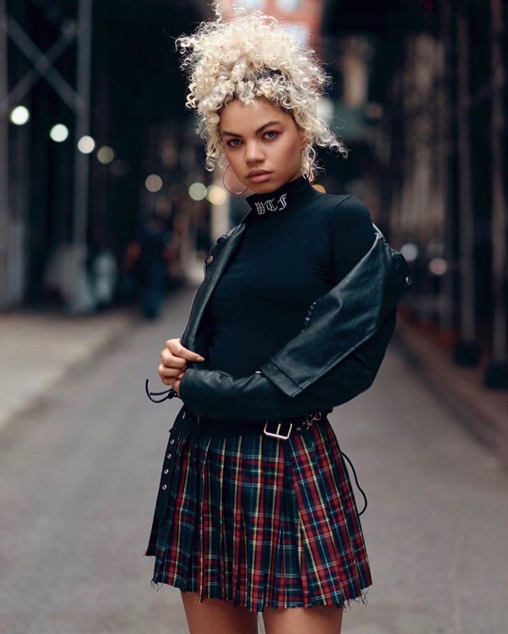 a young woman with curly blonde hair, styled in a high bun, stands confidently in the middle of a city street. she is wearing a black turtleneck, a red and green plaid mini skirt, and a black leather jacket draped over her shoulders. the background features blurred buildings and city lights, creating a sense of urban atmosphere. the lighting is soft and natural, likely from an overcast sky.