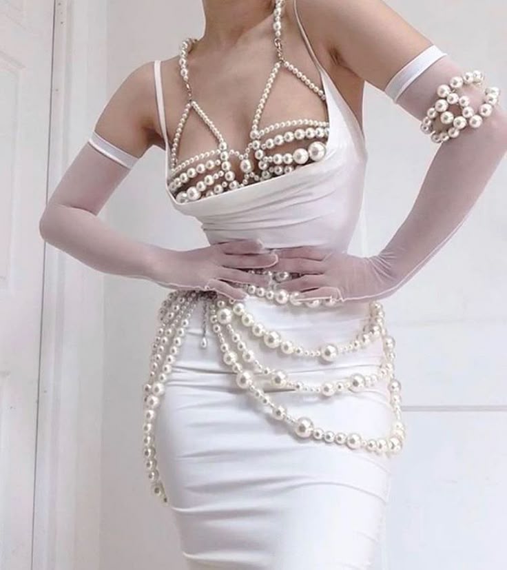 a woman is wearing a white satin dress adorned with various pearl jewelry. she has a pearl bra, a layered pearl body chain, a pearl bracelet on her arm covered by a sheer glove, and a pearl necklace. she has sheer gloves on. her hands are resting on her waist. the background is a plain white wall.