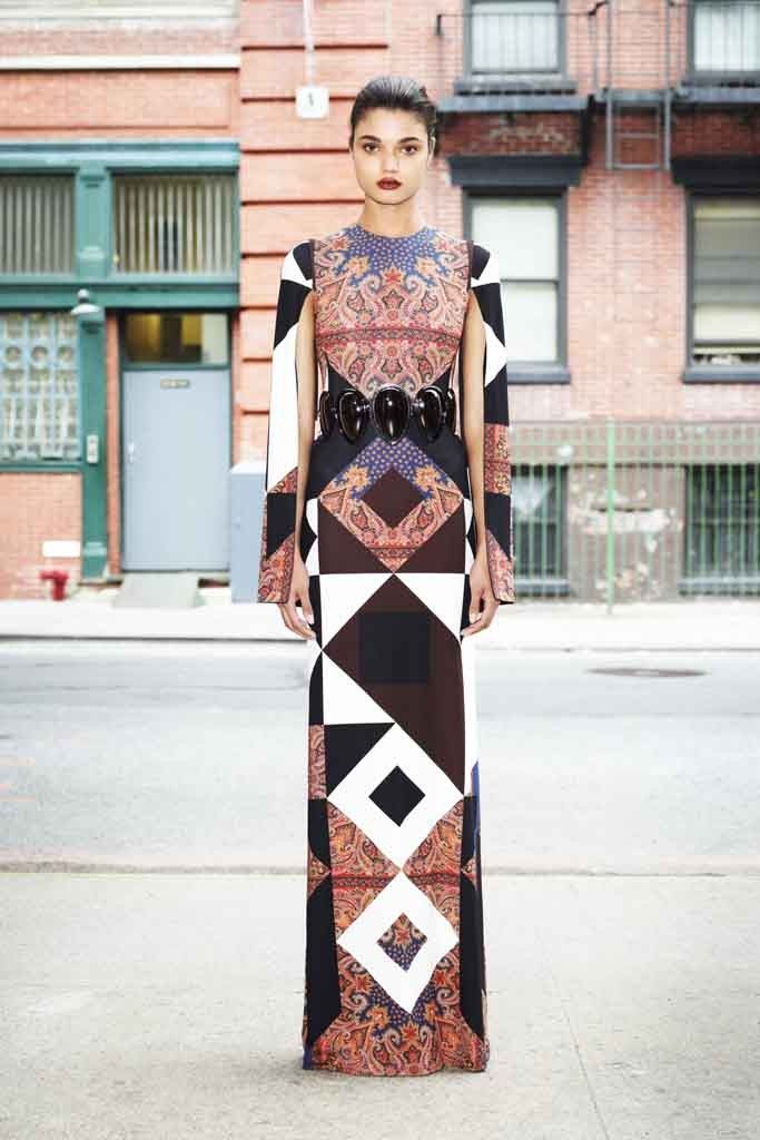 a fulllength portrait of a woman standing on a city street wearing a long, patterned dress. the dress features a mix of geometric shapes and paisley patterns in various colors including black, white, brown, and blue. the dress has a large black belt with three large circular embellishments. the woman has her hair pulled back and is looking directly at the camera. the background shows a cityscape with brick buildings and street features. the image is taken outdoors during the day.