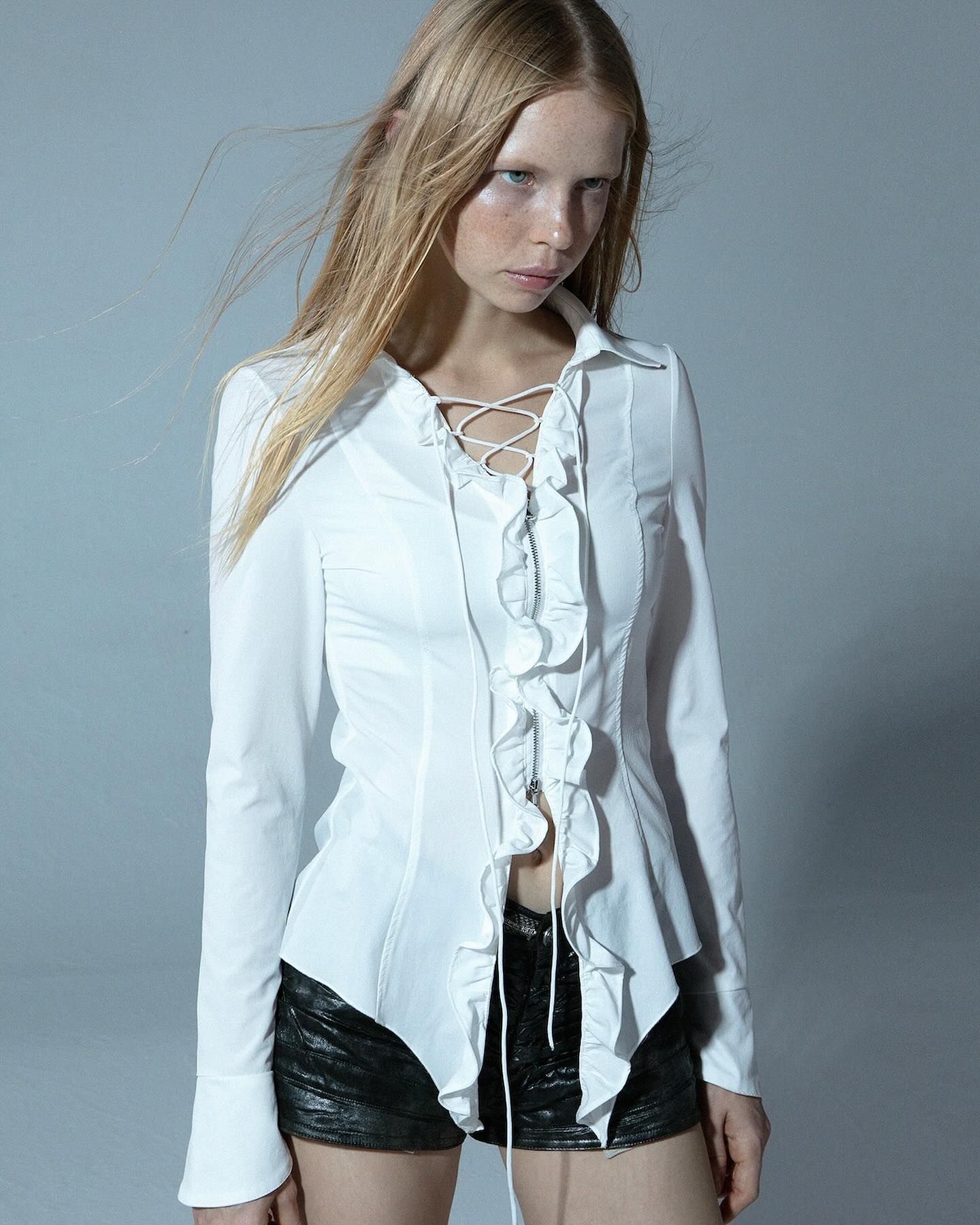 the image shows a slender blonde model wearing a stylish white longsleeved blouse. the blouse is fitted, featuring a laceup front and prominent ruffles cascading down the center.  the model is wearing black leather shorts. she is posed against a light grey background in a studio setting. the overall style could be described as high fashion, with hints of gothic or alternative influences. the focus is on the details of the blouse and the model's pose.