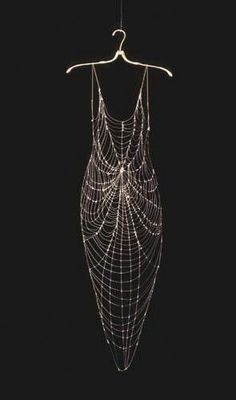 a silver spiderweb dress hangs on a hanger in a dark studio. the dress is made of  a delicate weblike pattern that creates the illusion of a spiderweb.  the dress is shimmering and sparkling, creating a luxurious and ethereal effect.  the dress's  halter neck style creates a chic and elegant feel. this is a fashion art piece that combines beauty and vulnerability in a stunning way.  the dark background  emphasizes the dress's delicate nature.