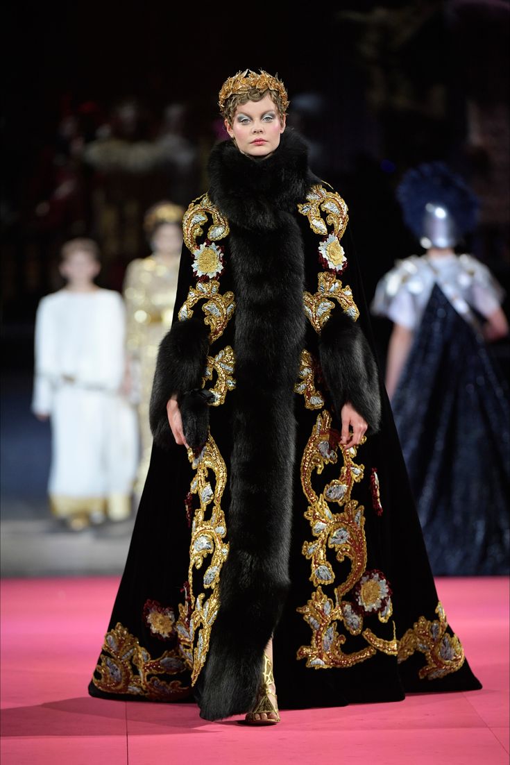 a model walks the runway in a stunning dolce & gabbana haute couture gown. the black velvet floorlength dress is richly embellished with gold embroidery depicting intricate floral patterns and swirling designs.  a luxurious black fur trim accentuates the neckline and edges of the capelike sleeves.  the model wears a gold crown, adding to the regal and majestic feel of the outfit.  the background shows other models and spectators in a blurred fashion show setting.  the overall impression is one of opulence and high fashion.