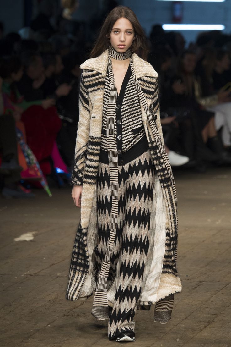 a model walks down a runway wearing a long, patterned coat and pants. the coat is primarily beige and black with a zigzag pattern. the pants match the pattern on the coat.  the model also wears a scarf around her neck. the background is blurred, showing the audience and a runway setting.  the overall style is contemporary and stylish.