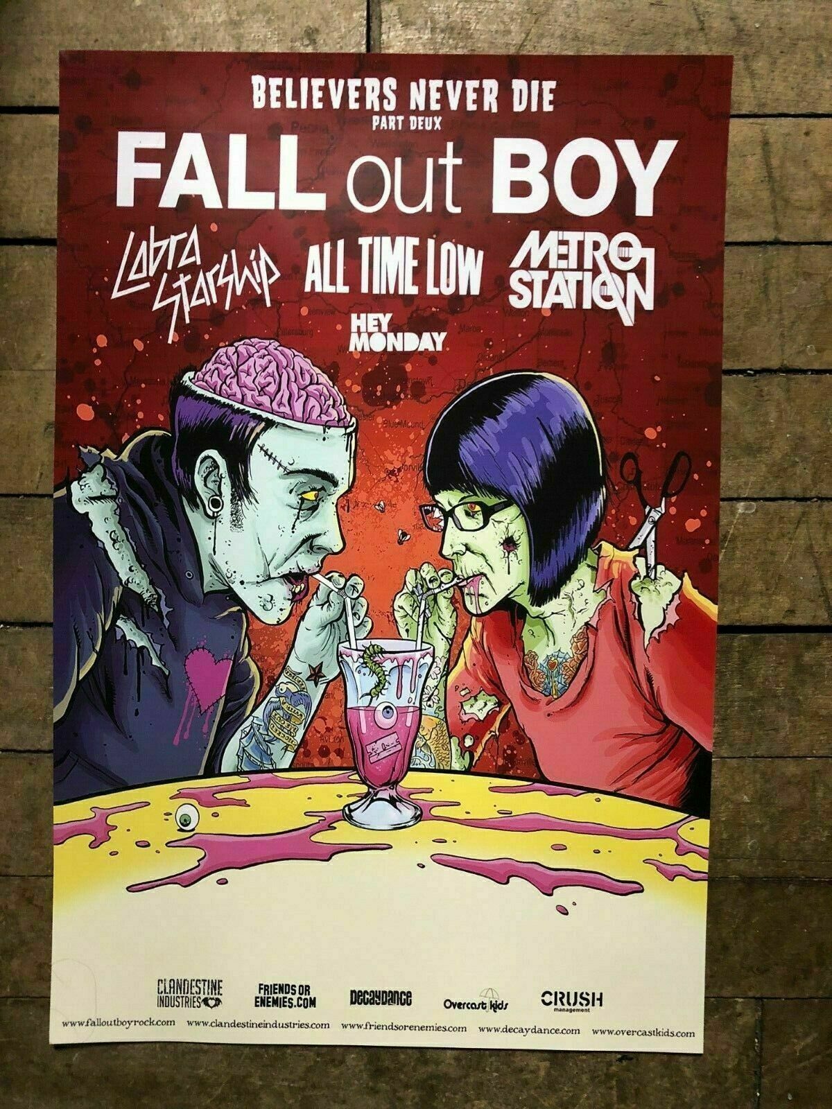 this is a concert poster for fall out boy's believers never die tour. the poster features a stylized illustration of two zombielike characters, one male and one female, sitting at a table and drinking milkshakes. the band's name is prominently displayed at the top of the poster, along with the names of the other bands on the tour. the poster also includes the tour dates, ticket information, and the web addresses of the bands and tour organizers.