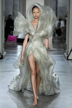 a woman model walks down a runway wearing a dramatic and ethereal silver gown. the dress is made of a sheer, flowing fabric that creates a billowing effect, creating a dramatic and elegant look. the model is posed in a fashionforward way, her legs long and slender, emphasizing the gown's design. the overall impression is one of high fashion and sophistication.