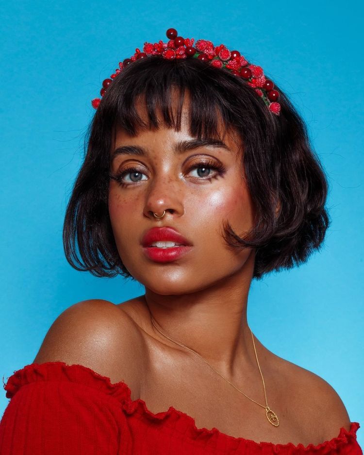 a closeup portrait of a young woman with brown skin and freckles. she's wearing a red offtheshoulder dress and a red floral crown, which looks like it might be made of berries. her makeup is subtly defined, and she has a septum piercing and a gold necklace. her short bob haircut features bangs, framing her face. the background is a solid, vibrant blue. the overall aesthetic is stylish, elegant, and youthful. the image appears to be a professionally taken studio shot.
